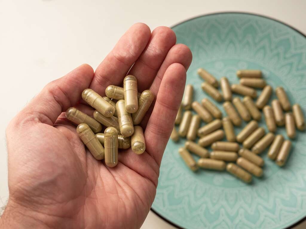 Ruka držiaca kratomové kapsuly a ďalšie kapsuly na tyrkysovom tanieri